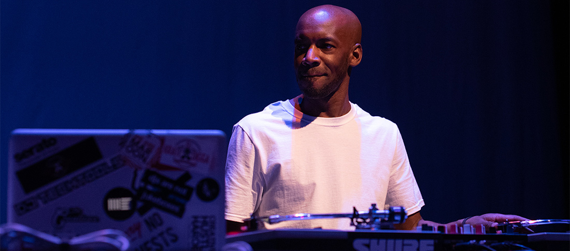 A black man with a clean shaven head, wearing a white t-shirt, stands in a mostly darkened space on a stage lit by blue lights. Two turntables and a laptop are placed in front of him. He looks to his right with a slight smile as he rests his left hand on one of the turntables.