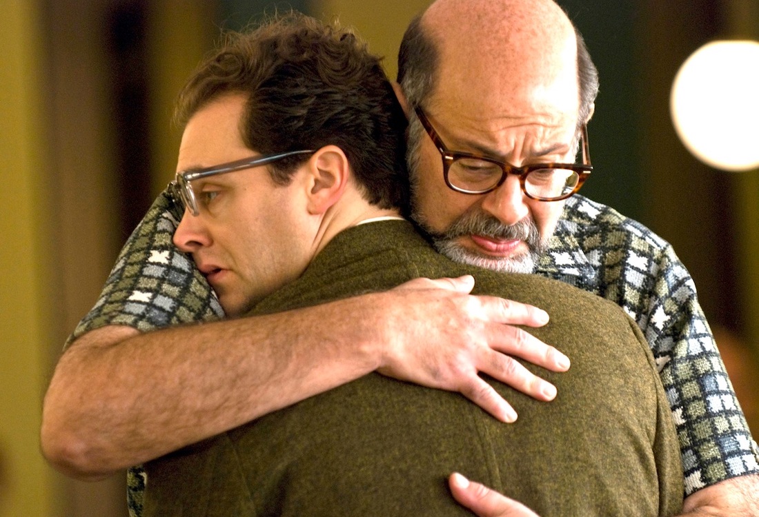 Film still of two white, middle-aged men in a tight embrace. The man facing the camera is balding and bespectacled with a solemn, downward gaze. The man he embraces is a little younger, with dark hair and glasses. He stares off to the side, looking dazed and uncomfortable.