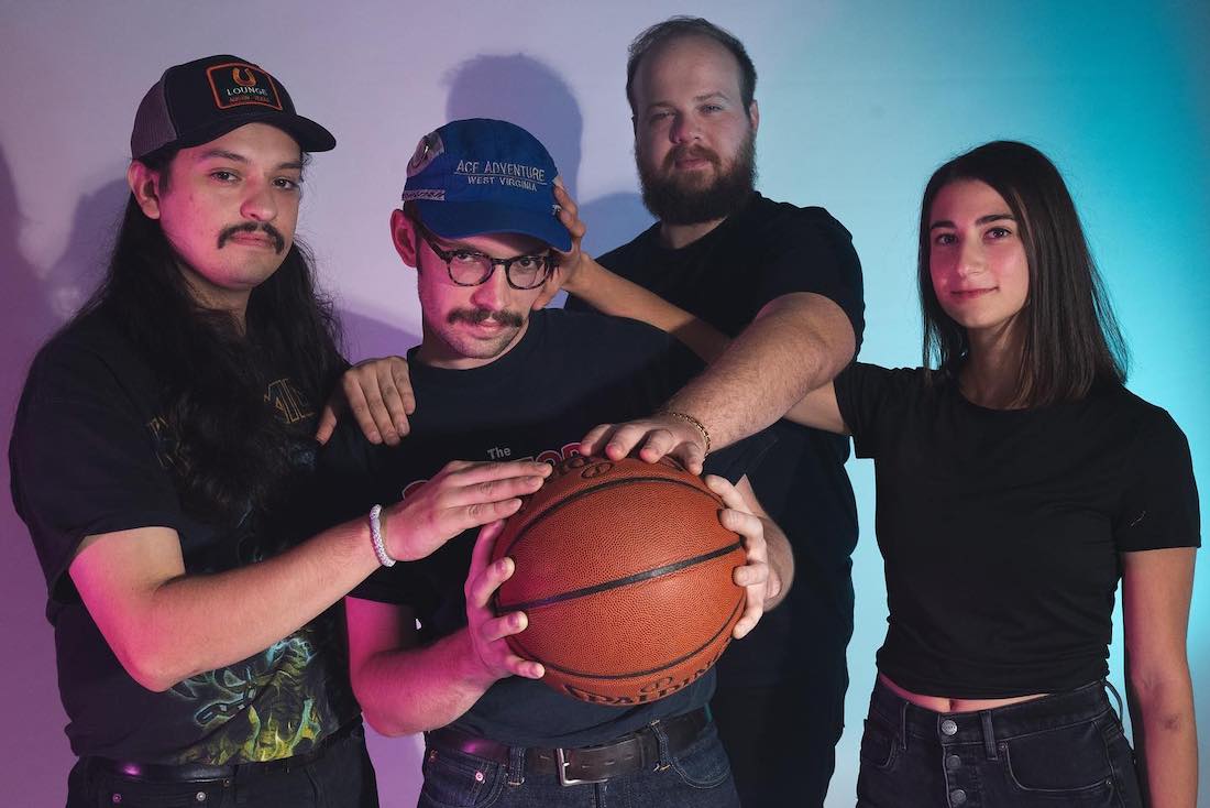 Three white men and a white woman stand close together in a space lit in blue and pink. One man stand near the center holding a basketball. The other two men stand on either side of him, each with one hand on the central man's shoulders and one hand on the basketball. The woman stands to the right with one hand placed on the central man's baseball hat-clad head.