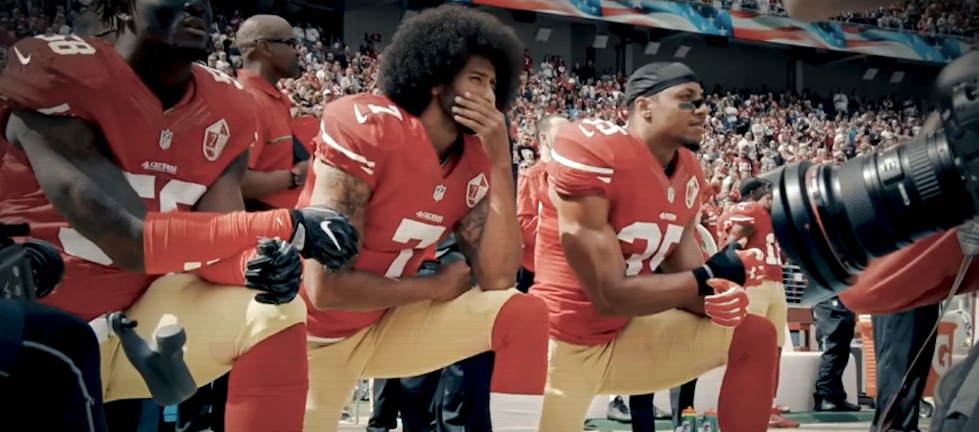 Three Black football players kneel on the sidelines of a game, staring forward intently. A crowd is in the stands behind them. A long telephoto lens is on the right of the frame, pointed at the players.