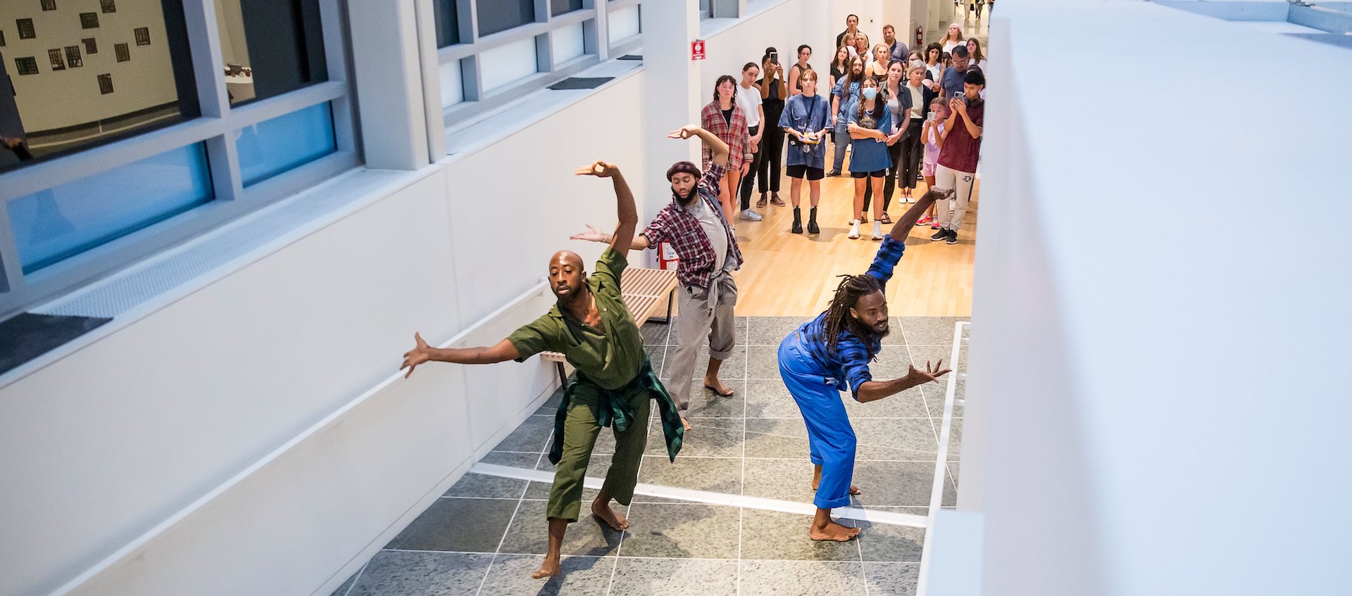 Three Black dancers performing up the Wexner Center gallery with their arms outstretched as a crowd follows them up the ramp.