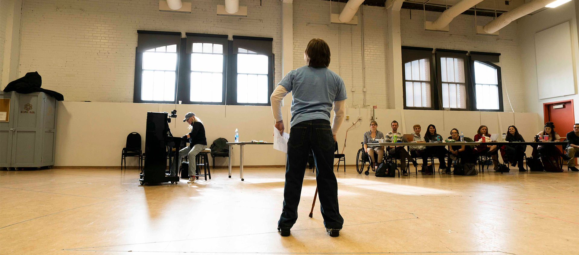 A person performs, cane in hand, in an auditorium. In the background, a person plays a piano next to a line of people sitting and watching the performer.