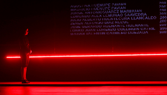 A person in all black stands in front of a long, red neon light running parallel to the floor. Illegible text is projected on the wall above it.