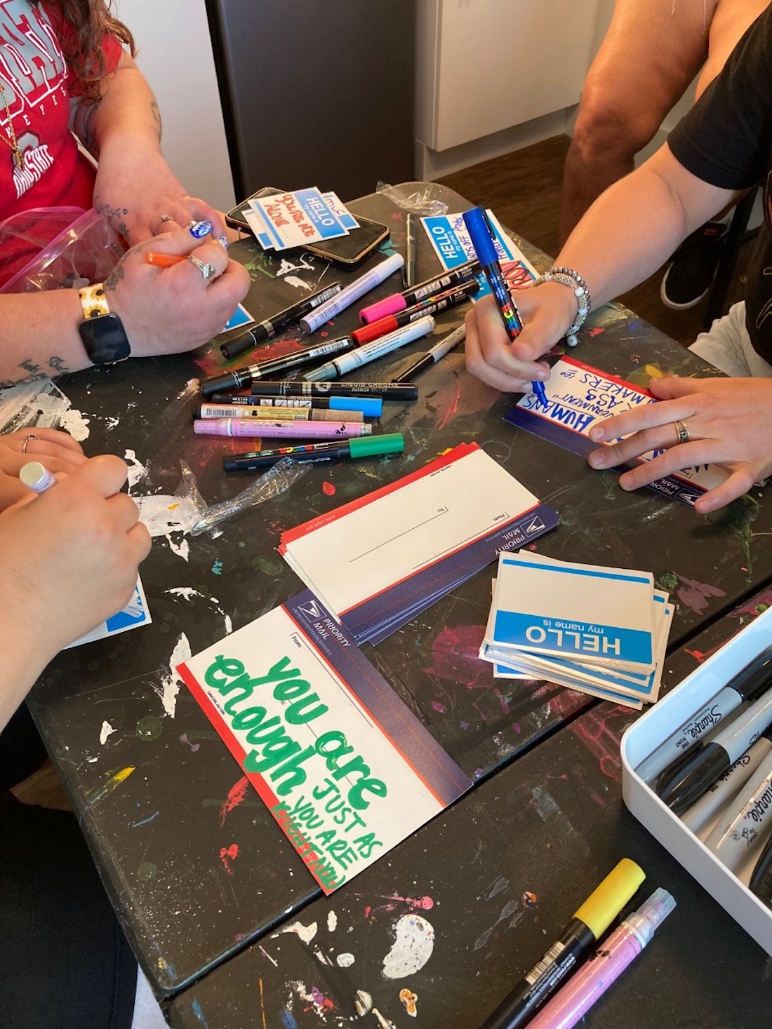 A paint-spattered work table holds markers and blank name tags. Individuals seen from shoulders down hold markers as they embellish the name tags. A small sign on the table reads, "You are enough just as you are."