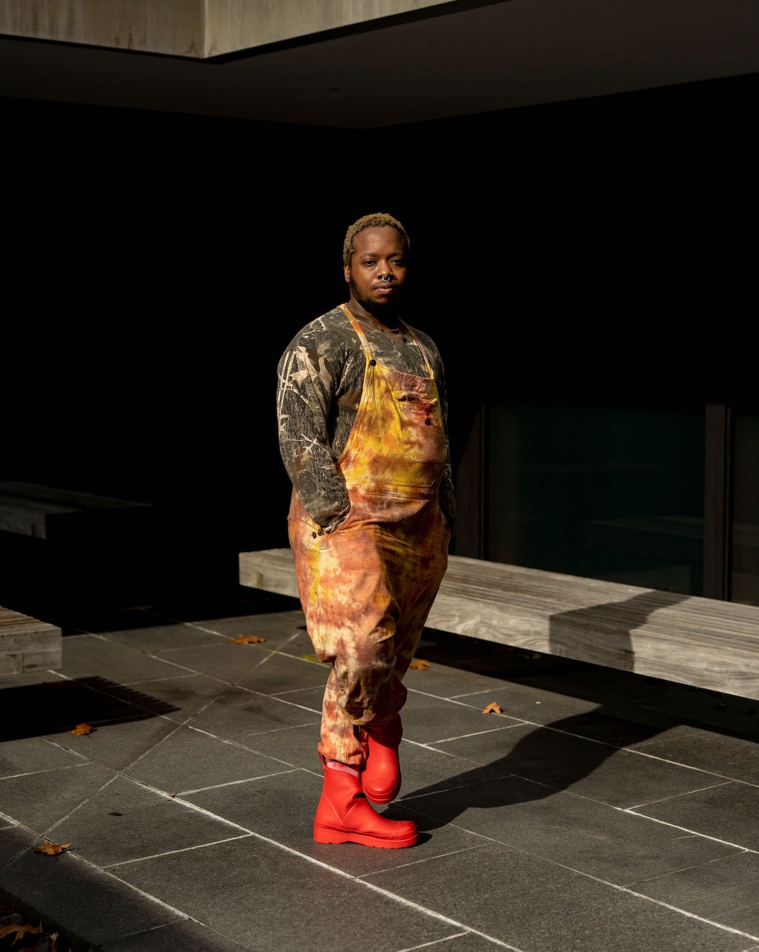A Black man stands at the center of a space with black walls and a gray stone tile floor. He has a short, blonde-dyed Afro and is wearing a paint-covered sweatshirt and overalls with new red ankle boots. He's turned slightly away from the camera but looks directly at the viewer.