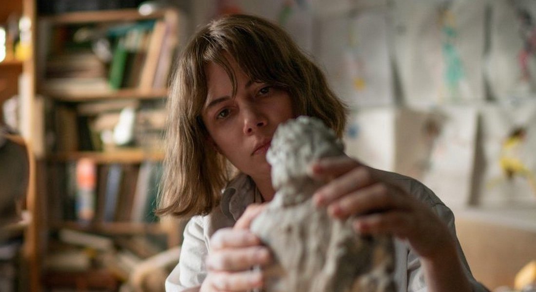 A white woman with shoulder length brown hair with bangs is shot shoulders-up in a cluttered artist studio. She's intently concentrated on shaping a small figurative sculpture in clay.