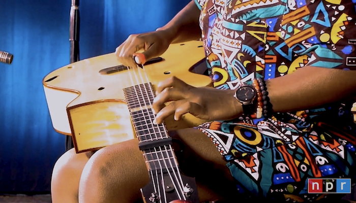 Video still of guitarist Yasmin Williams sitting, shown only from chest to knees, with a shadowy, deep blue wall in the background. Williams is wearing a top and matching shorts in a bright, multicolored African print. Williams has an acoustic guitar on her lap and she's picking its strings with both hands.