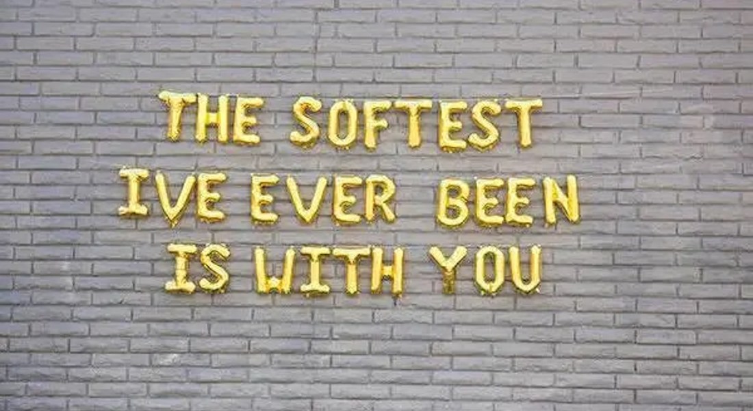 The words "The softest I've ever been is with you" are spelled out in gold letter balloons, placed against a bric w