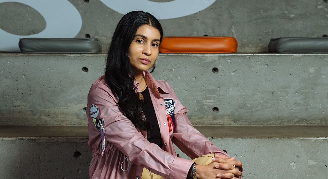 A Muslim-American woman with long, straight black hair, sits against a concrete wall. She wears a black t-shirt with a pink, patch-covered collared shirt over it. Her body is turned to the left but her face is turned toward the camera, with a direct gaze.