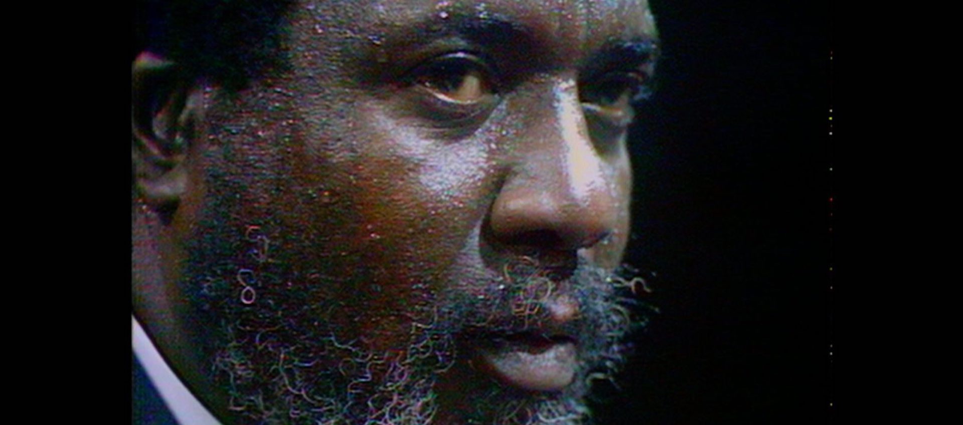 Tight close-up of the face of a Black Man with short hair and a gray-flecked beard. He is looking off to his left.