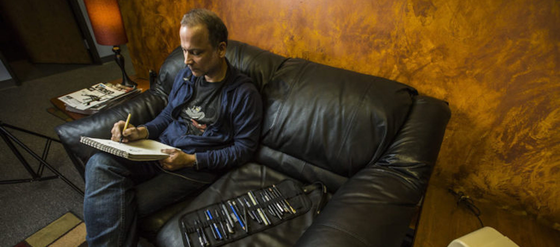 A man sits on a leather loveseat in a warmly lit living space, sketching in a sketchbook. A large, filled sleeve for drawing tools sits open beside him