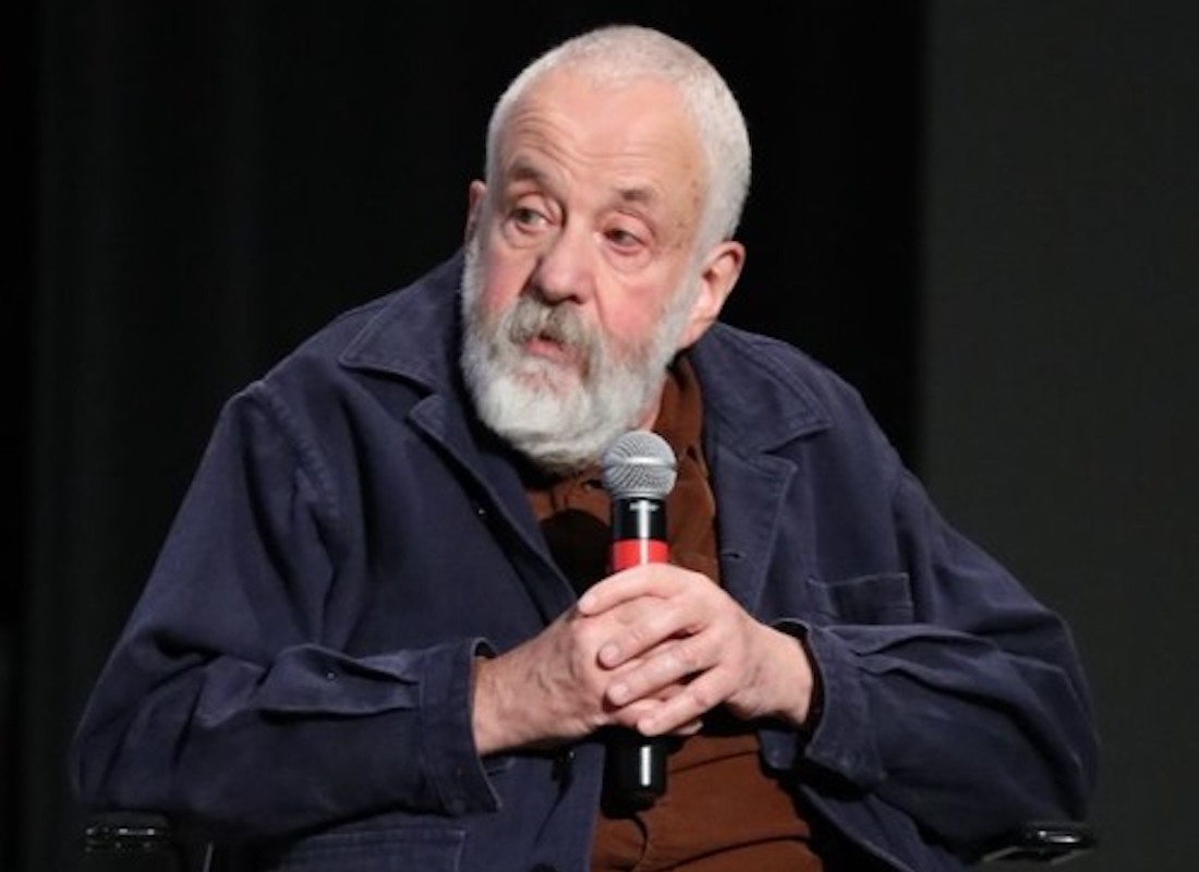 A man with white thinning hair and a white bear sits on a darkened stage, holding a microphone with both hands.