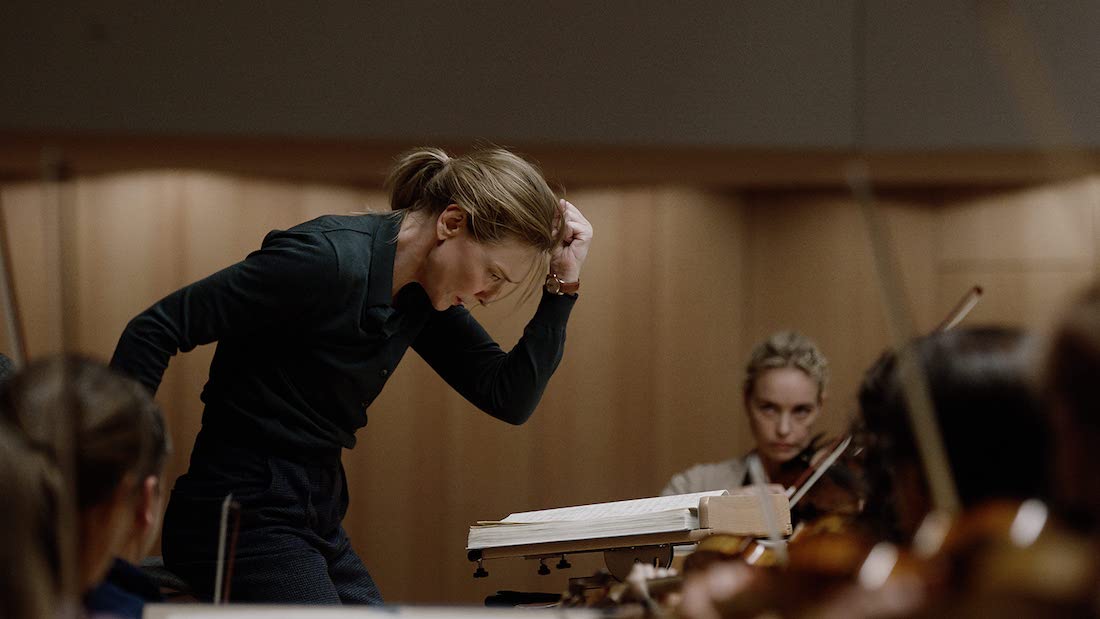 A woman dressed in dark clothing, captured from the side and from the hips up. She is in a rehearsal space and is caught mid-motion while staring intently at the score on a podium in front of her. A few musicians can also be seen in front of her. All are staring at her.