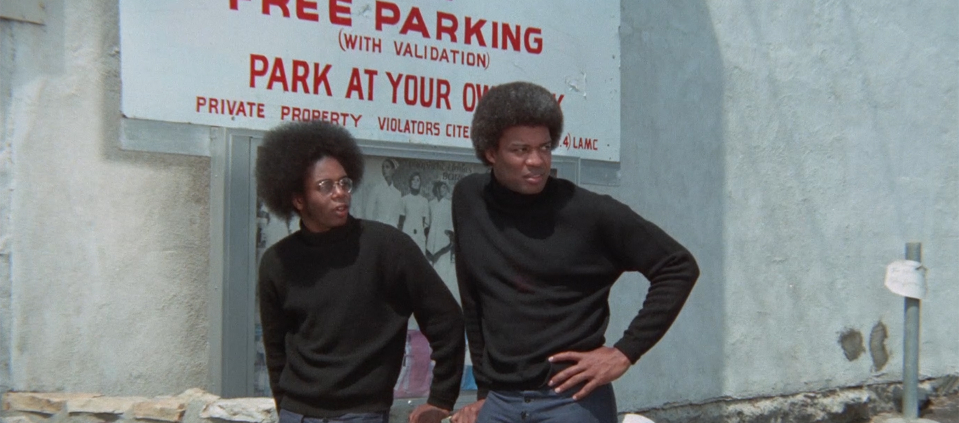 Two Black men stand in front of a parking lot sign, looking to the right, and wear black, long-sleeved turtlenecks and their hair in afros.