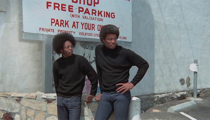 Two Black men stand in front of a parking lot sign, looking to the right, and wear black, long-sleeved turtlenecks and their hair in afros.