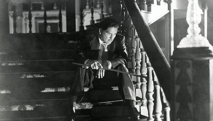 Black-and-white still of a man in a suit. He sits on a staircase, holding a cane and looking downward, as if in deep thought.