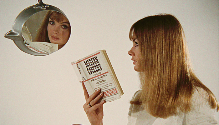 The profile of a white woman with long, brownish blonde hair. She holds a book titled Modern Cookery and looks at her reflection in the bottom of a frying pan hanging above her.