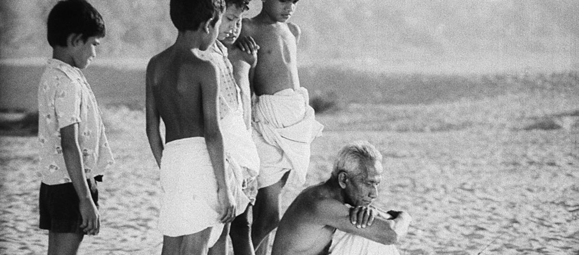 Black-and-white still of a group of Indian people on the beach, including five standing children and an older man who sits in front of them with his arms crossed on his knees.