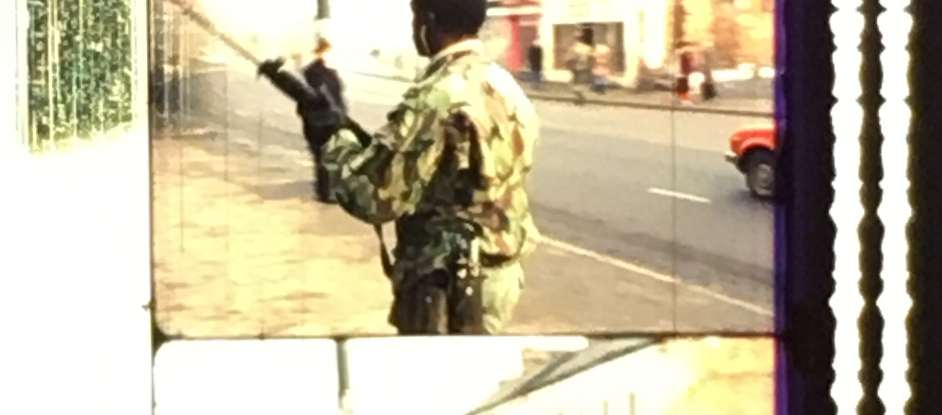 A strip of frame enlargements from a film print. The image shows a Black man from behind who is dressed in military fatigues. He is holding a rifle and standing in a city street.