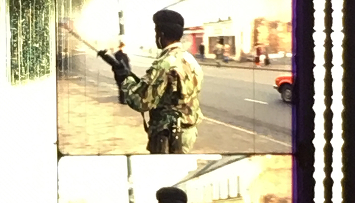 A strip of frame enlargements from a film print. The image shows a Black man from behind who is dressed in military fatigues. He is holding a rifle and standing in a city street.