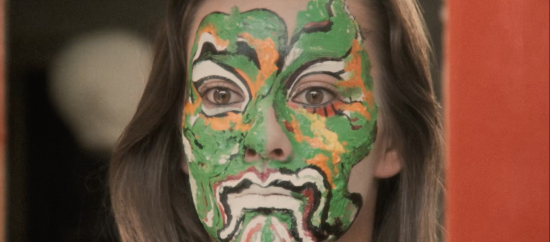 A close-up still of a woman with her face painted, in a pattern of green, orange, white, black, red, and yellow, to look like an ornate mask.