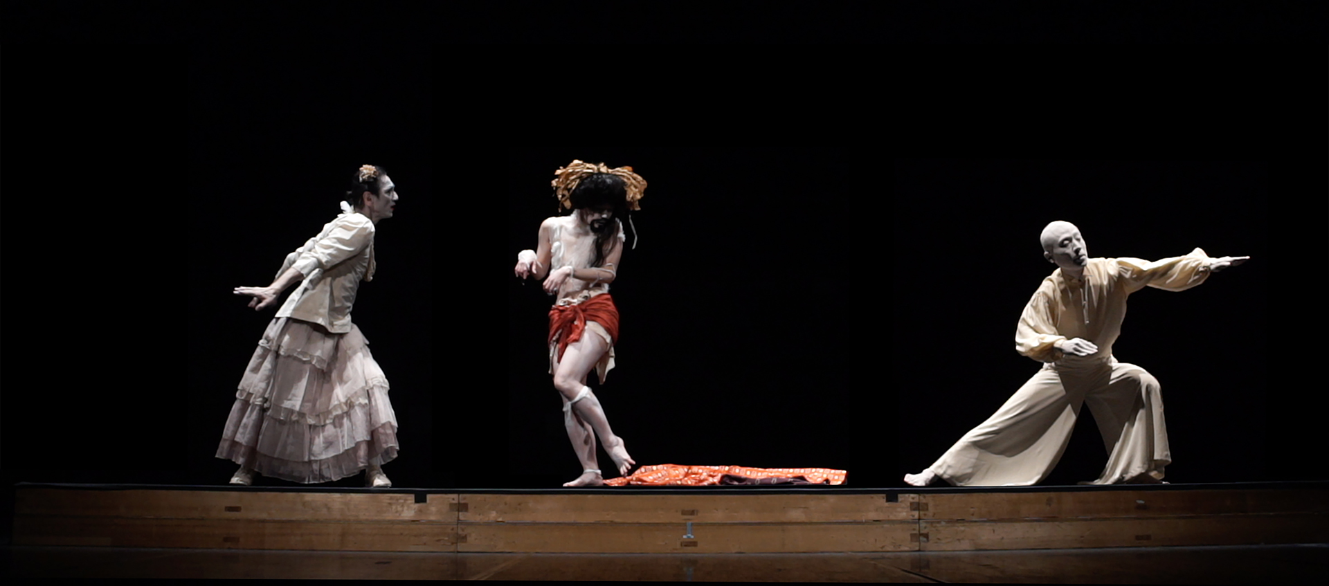 Three Butoh dancers are shown on a stage from a distance against a black background. Their skin is painted with white makeup. 
