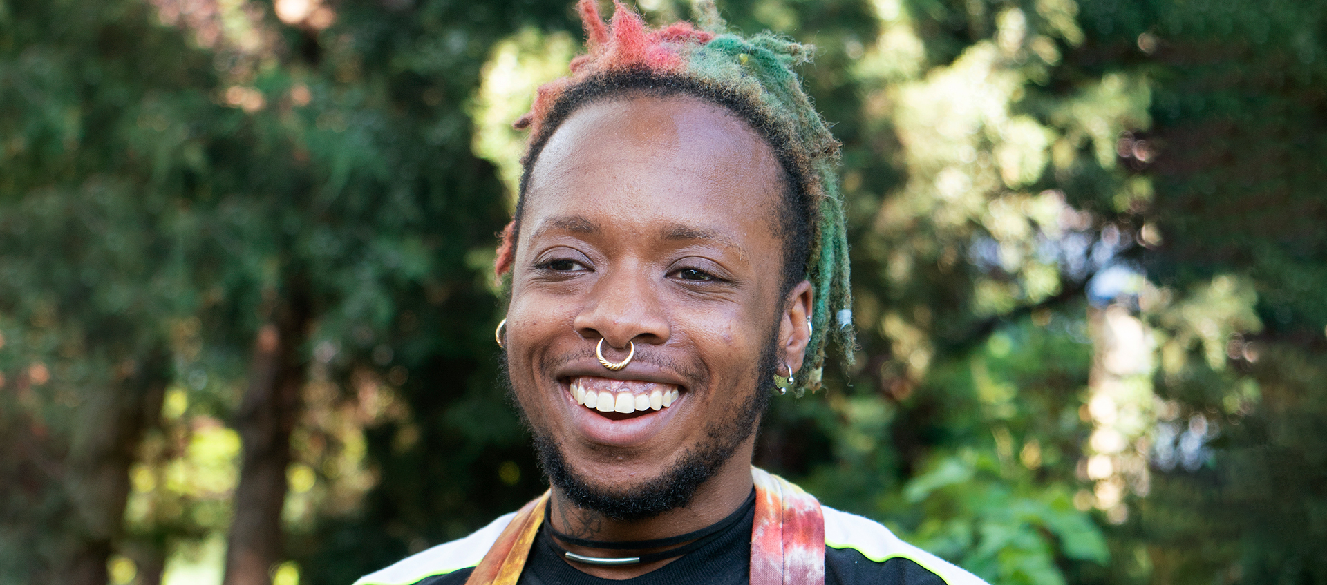 Anaïs Duplan smiles against a backdrop of trees. He has medium brown skin; short, red and green locs; black facial hair; and a septum and ear piercings.