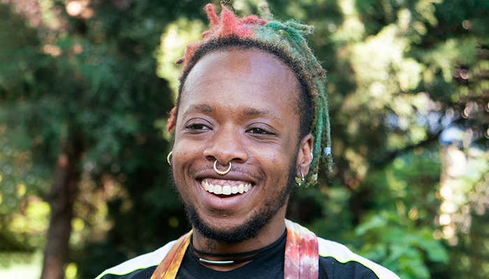 Anaïs Duplan smiles against a backdrop of trees. He has medium brown skin; short, red and green locs; black facial hair; and a septum and ear piercings.