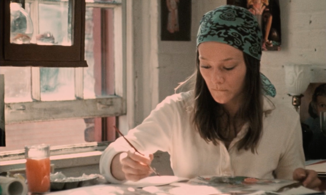 A woman is sitting at a table painting. She is wearing a light top and has a scarf wrapped around her head. 