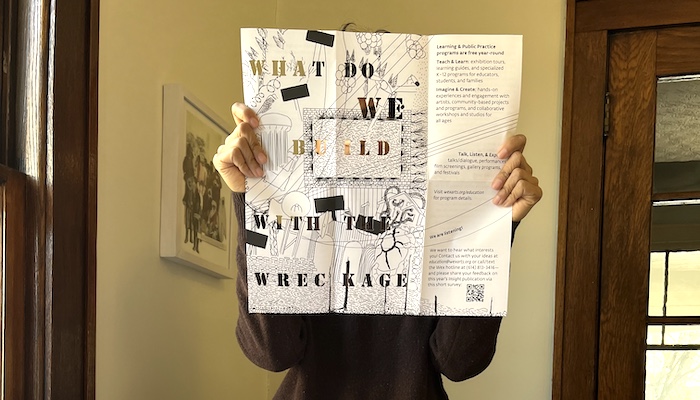 Standing inside a warmly lit living space, a person holds in front of their face a black-and-white foldout brochure with cutout text