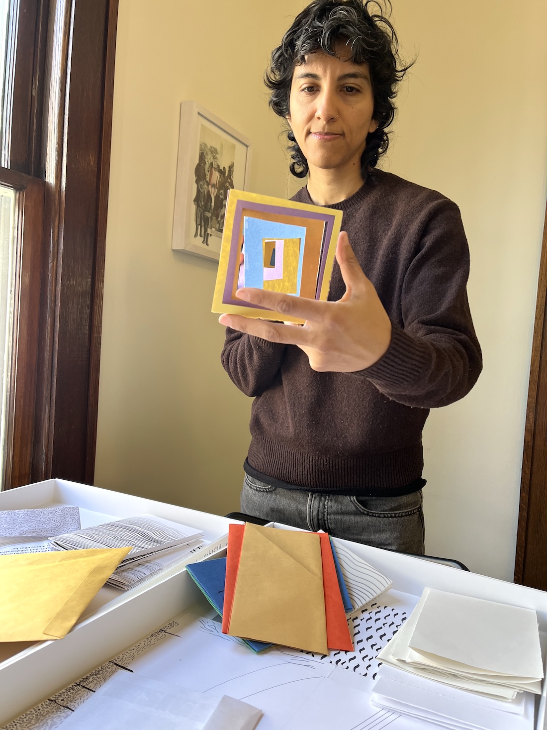 A person stands in a warmly lit living space with a table littered with construction paper and art supplies. They hold a small, colorful paper collage.