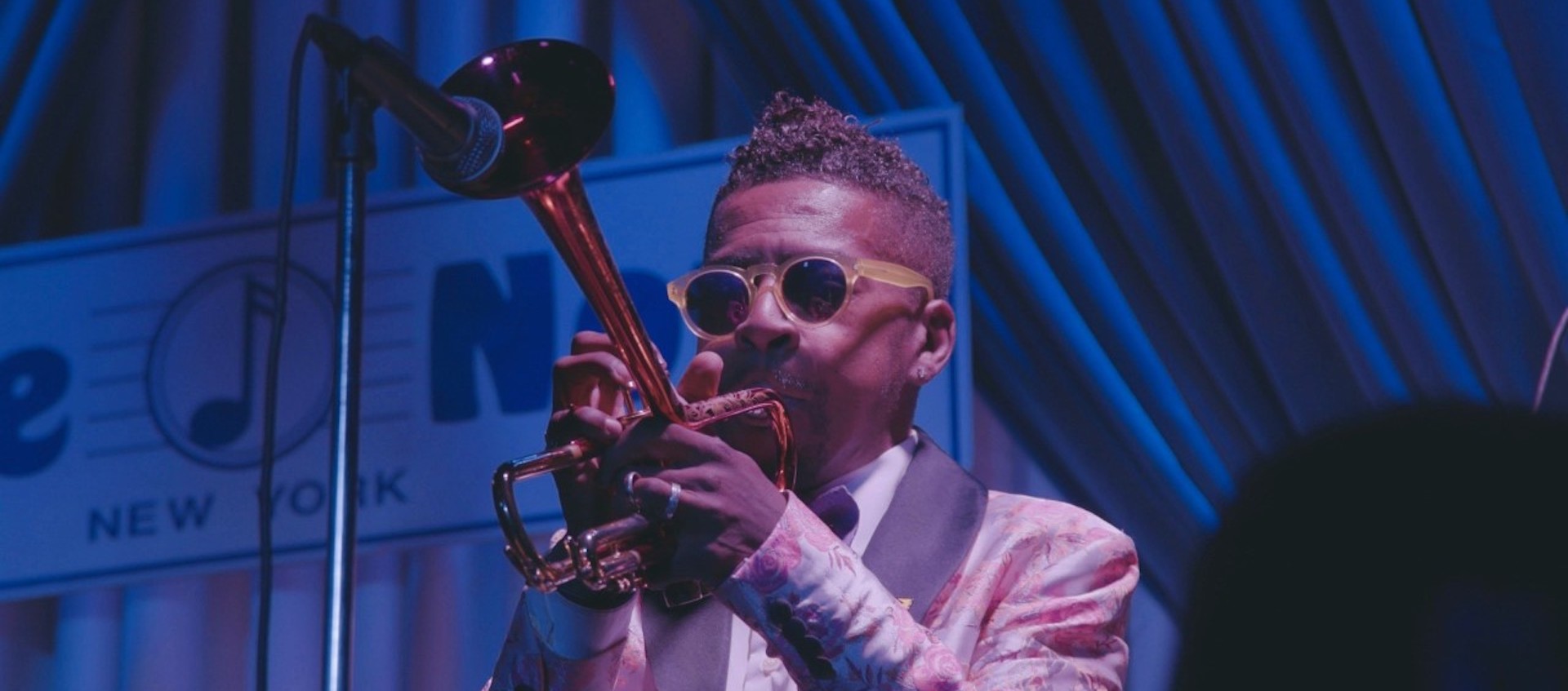 A trumpeter wearing dark glasses and a floral patterned tux plays his instrument into a microphone on a blue-lit stage.