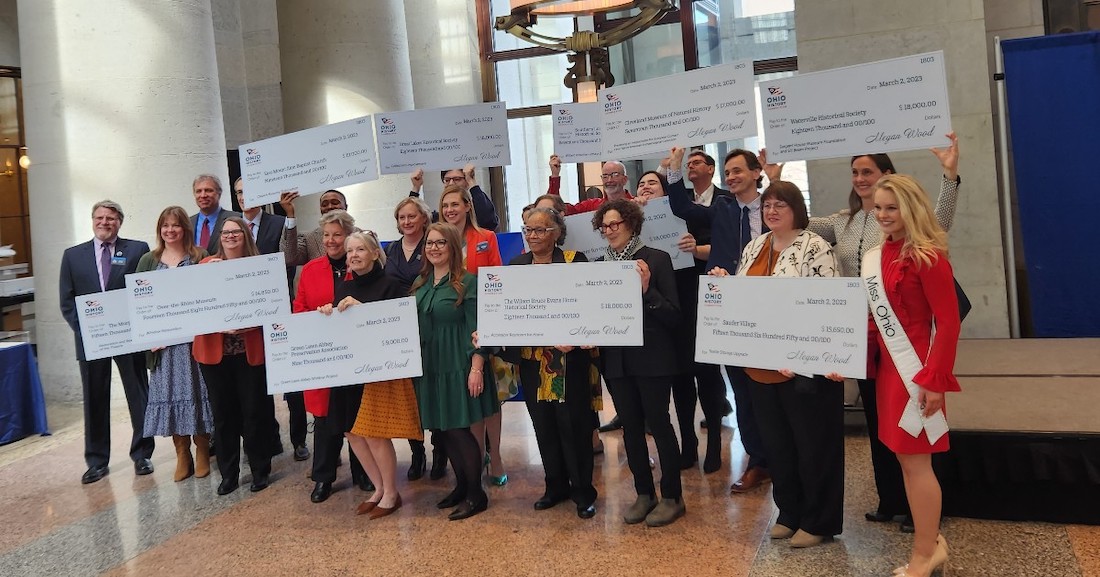 About 20 individuals stand in the hallway of the Ohio Statehouse. They are gathered close and smiling. About 10 of them are holding oversized novelty checks indicating grant amounts won from the state historical fund.