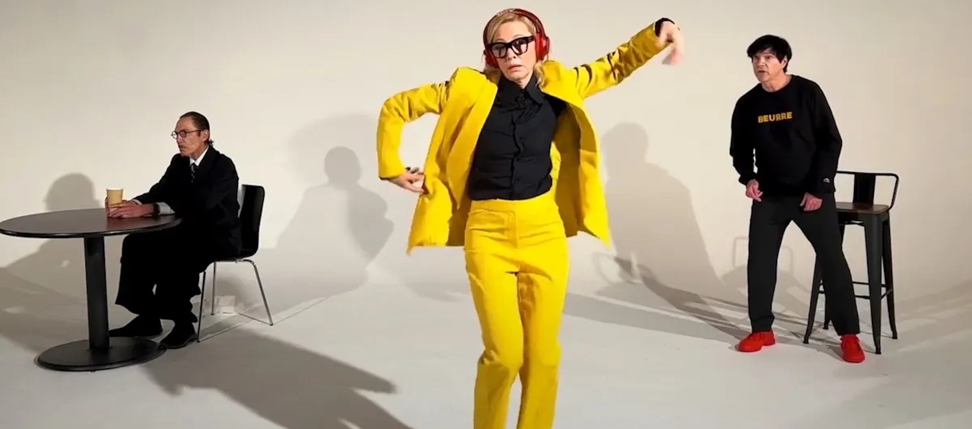 A woman wearing black-rimmed glasses, red headphones, and a bright yellow suit with a black shirt dances at the center of an all-white space. Behind her, a man to the right leans forward, singing, and a man to the left sits at a round black table, looking off in the distance. His hands hold a cup of coffee.