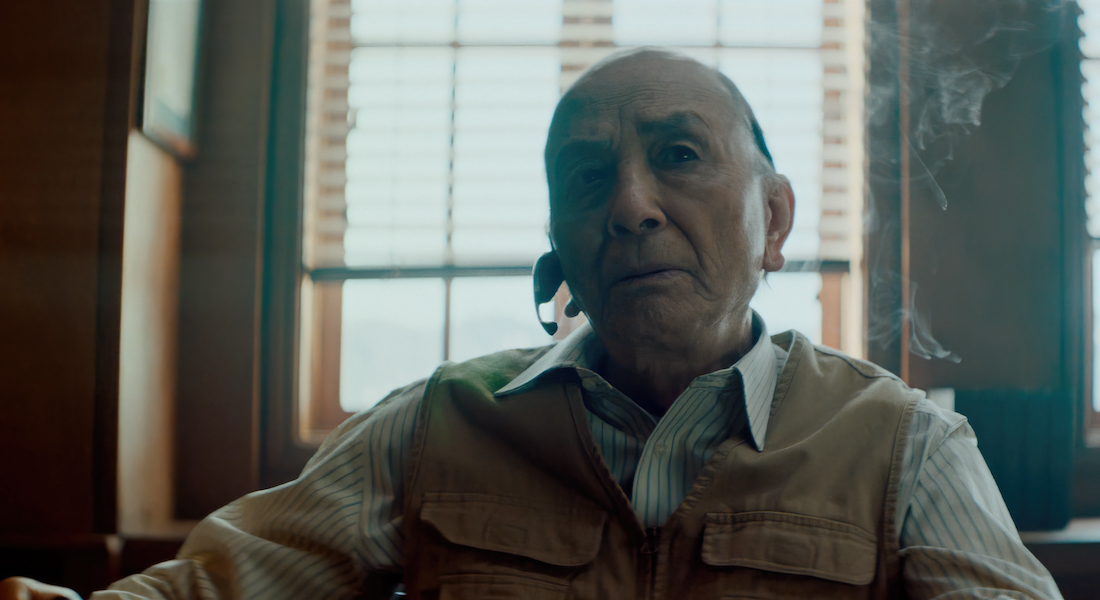 Close-up of an elderly man, shot from the shoulders up. He is wearing a striped shirt and khaki vest with pockets. He’s sitting in an office space and staring slightly to the left with a tired look in his eyes. Smoke from a cigarette rises in the air to his left.