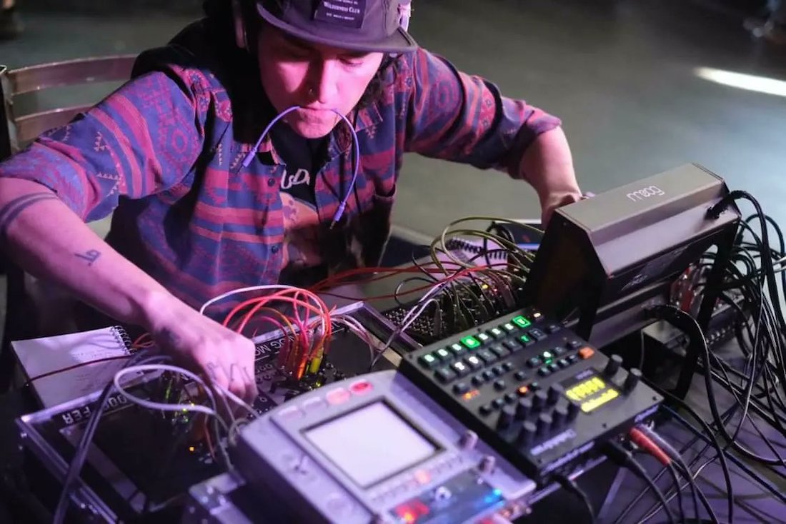 A person looks down at a synthesizer control board dense with patch cables. They’re clenching another patch cable in their mouth. They’re using two hands to adjust knobs on the board.