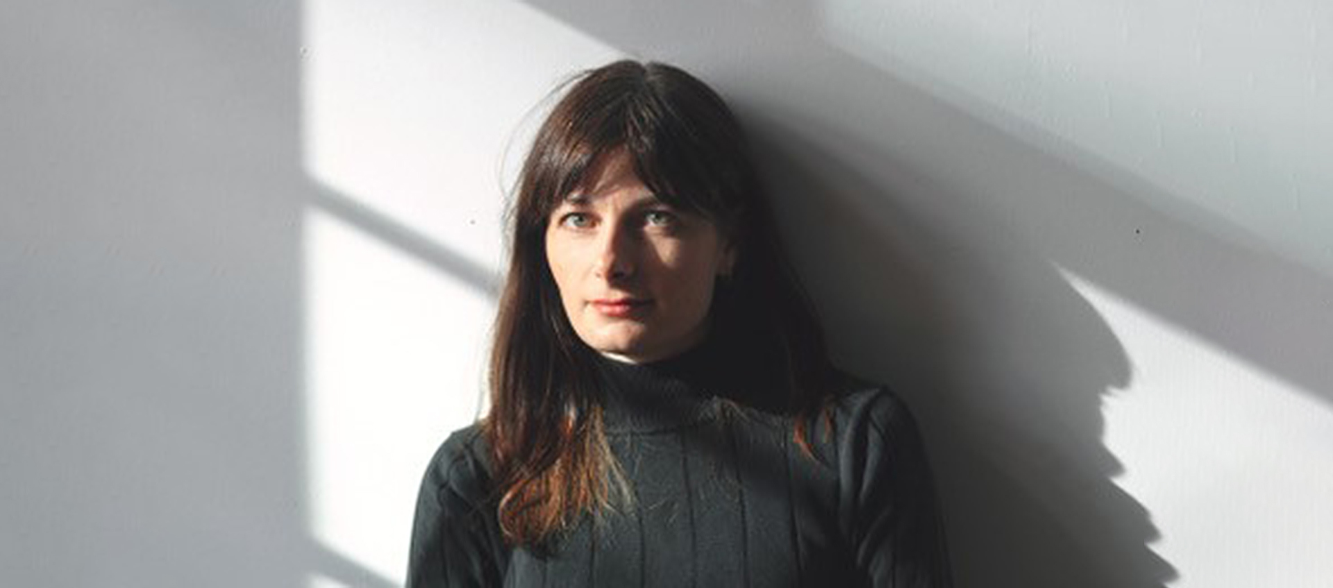 Brett Story, a white woman with light brown hair, sits behind a wooden table in front of a white wall. She is wearing a black turtleneck and the sun streams in through some off-frame windows, creating dynamic shadows and patches of light around her. 