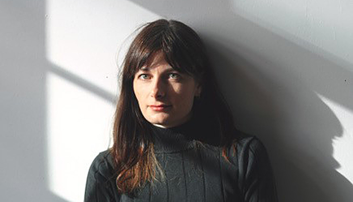 Brett Story, a white woman with light brown hair, sits behind a wooden table in front of a white wall. She is wearing a black turtleneck and the sun streams in through some off-frame windows, creating dynamic shadows and patches of light around her. 