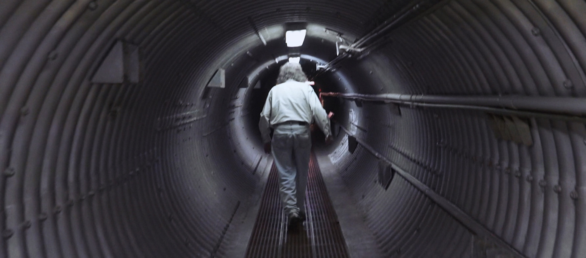 A gray-haired man with his back to the camera walks down a round metal underground tunnel.