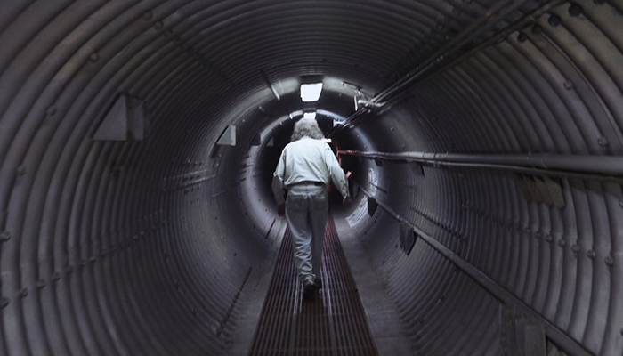 A gray-haired man with his back to the camera walks down a round metal underground tunnel.