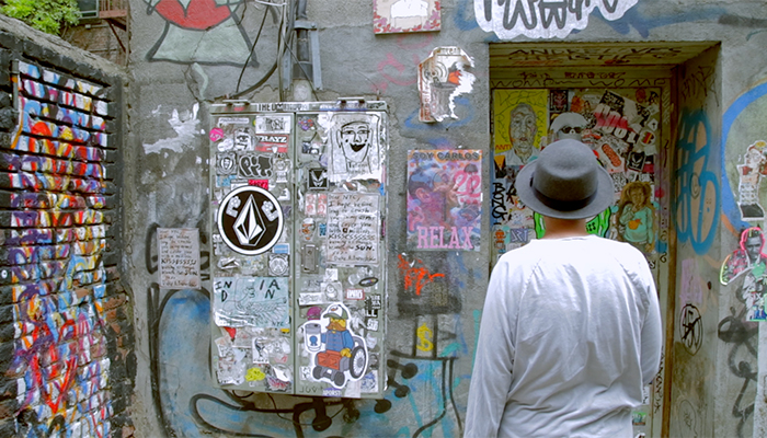 A man wearing a hat has his back to the camera while standing in an alley that’s covered with graffiti and flyers. The word “RELAX” appears prominently on one of the flyers.