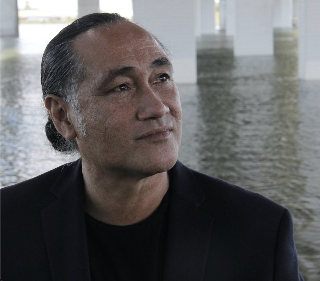 A man with long, dark, slightly graying hair that’s pulled back in a bun is shot from the shoulders up. He’s in a space with square white columns rising up from a body of water behind him. He’s in a dark suit with black t-shirt. He’s looking off in the distance to his left.