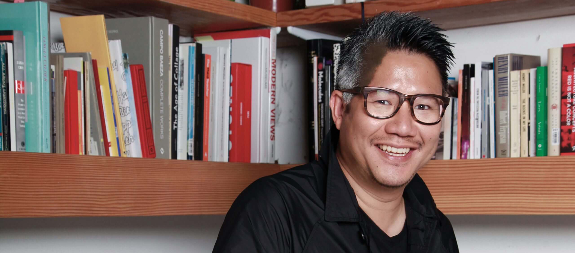 Kulapat Yantrasast, a man of Thai descent, stands in front of book shelves. He smiles at the camera, wearing a black shirt and glasses. He has short black-and-gray hair. 