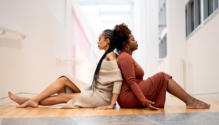 Two Black women sit on a gallery floor with their backs against one another. They are barefoot and have their eyes closed in meditation.