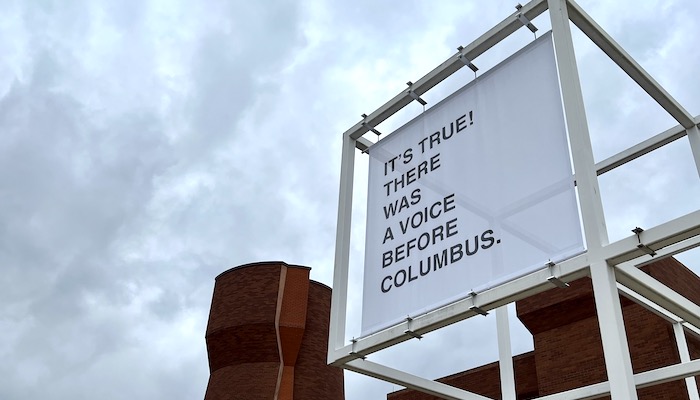 A large, white banner with black text reads, “IT’S TRUE! THERE WAS A VOICE BEFORE COLUMBUS.” It hangs outside within a white metal grid in front of a brick tower.