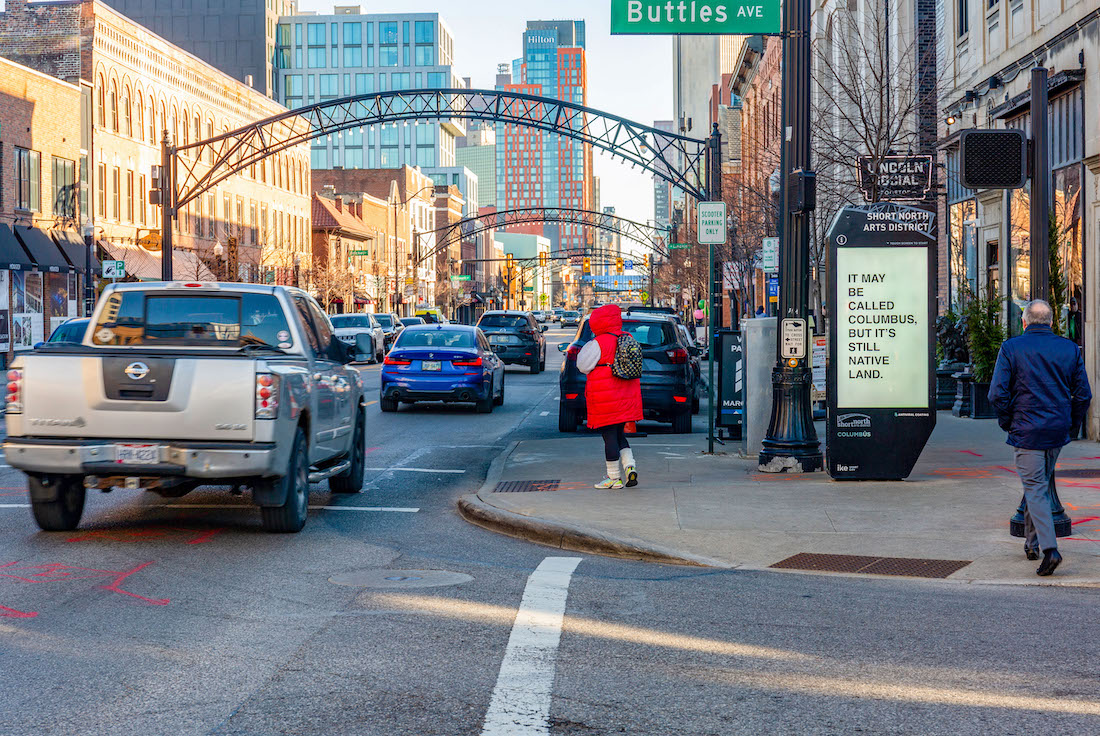 A long view of a street filled with cars. A row of vintage-style metal arches rise above it. On the right, a sidewalk corner holds a tall smart monitor with a video screen. The screen holds black text on a white background that reads, "IT MAY BE CALLED COLUMBUS, BUT IT'S STILL NATIVE LAND."