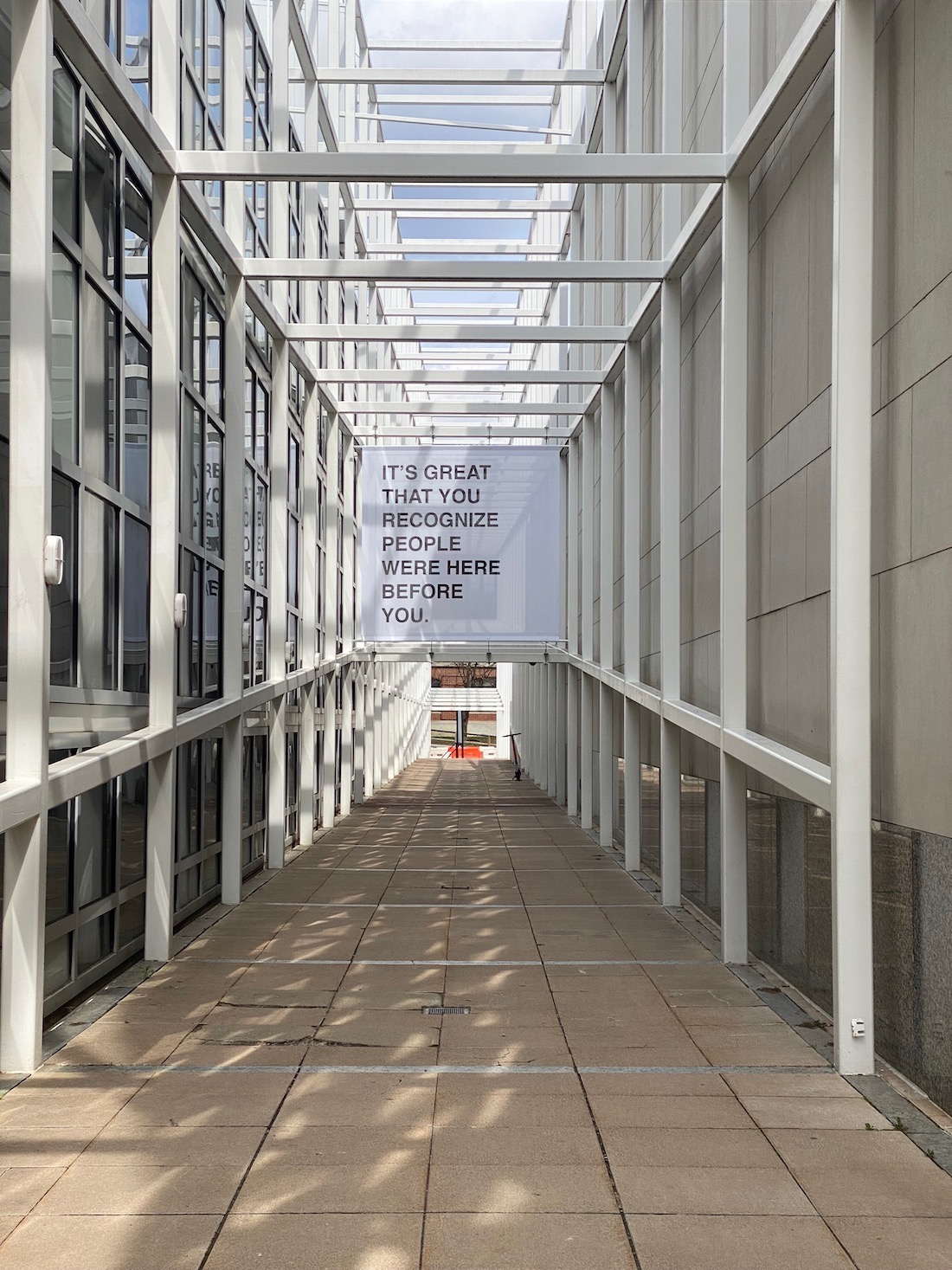 A walkway contained by an open grid of white metal bars, on which a text work is installed overhead. It reads, "IT'S GREAT THAT YOU RECOGNIZE PEOPLE WERE HERE BEFORE YOU"