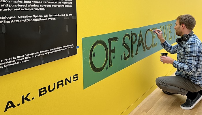 Using a stencil, an artist paints an exhibition title on a bright yellow wall