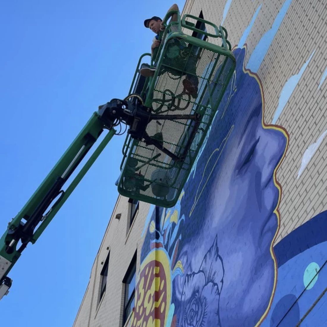 An artist stands on a cherry picker looking down. A light brick wall is behind him, adorned with a large portrait mural
