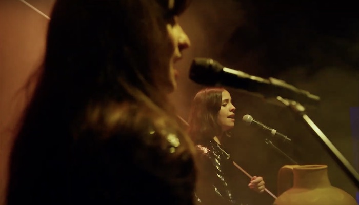 Close-up of two women singing into microphones on a dark stage touched by soft amber light